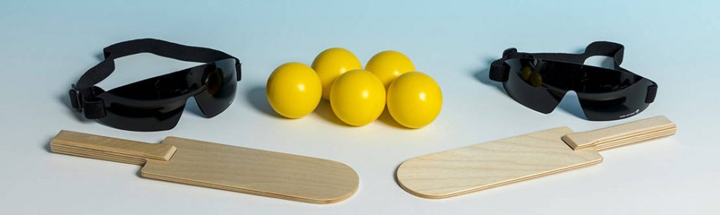Picture showing showdown equipment including two paddle like bats, two pairs of eye-shades and five yellow balls