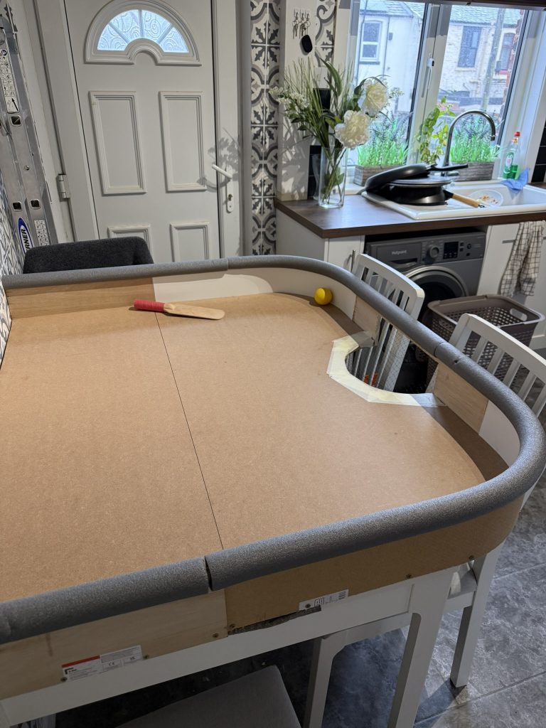 Photo of the home made practice table made from ply wood with side walls and curves leading to a goal pocket. on the table lies a showdown bat and ball.