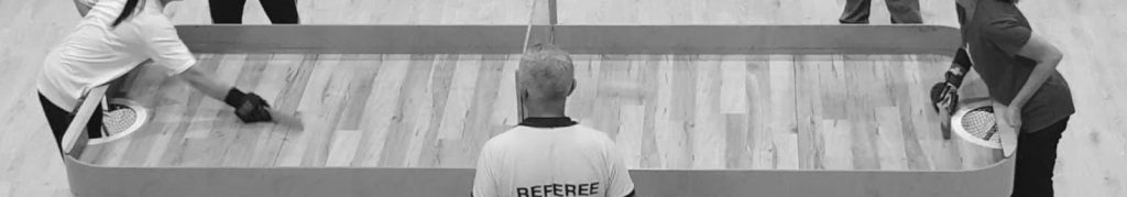 Black and white image of 2 people at either end of a showdown table in the middle of a rally, witha referee watching on.